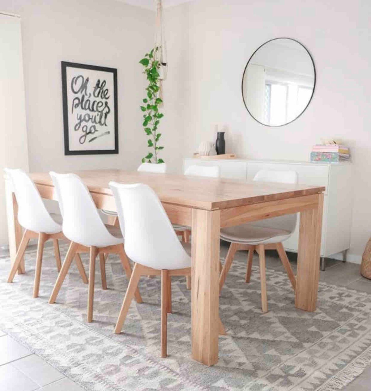 A light-filled dining area features a wooden table surrounded by five white chairs. A circular mirror hangs above a sideboard, complemented by a wall art piece with a welcoming phrase. A patterned area rug lies beneath, adding texture to the space.