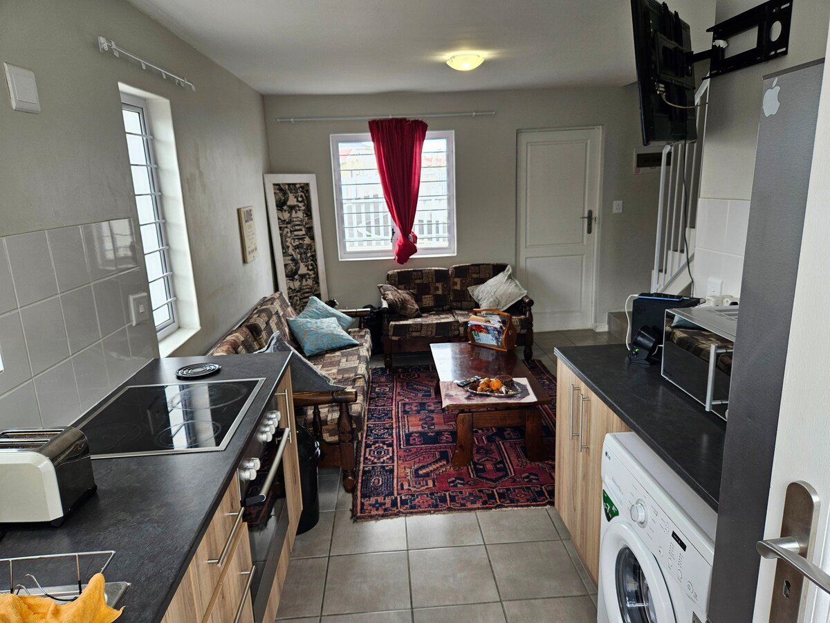 The living area features a combination of light-colored cabinets and dark countertops. A cozy seating arrangement includes a patterned sofa and armchair, complemented by a wooden coffee table. Natural light enters through two windows, and a flat-screen TV is mounted on the wall.