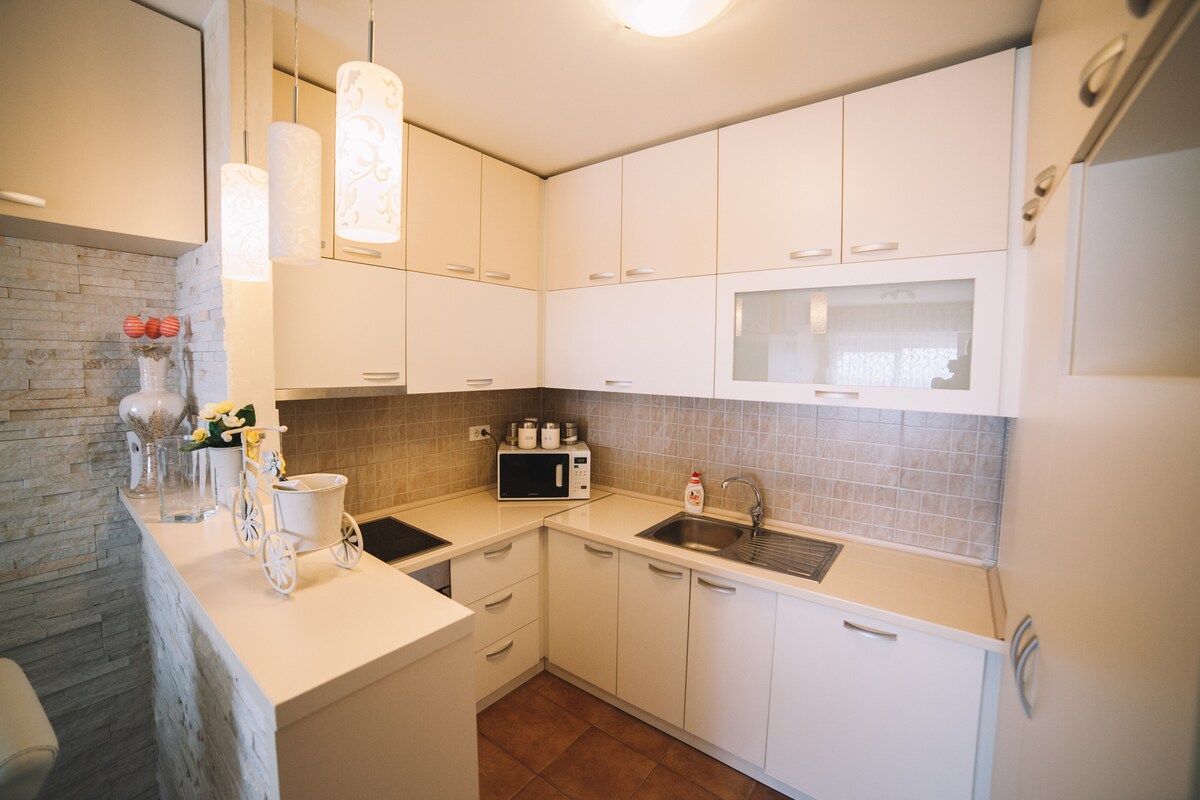 A modern kitchen features sleek white cabinetry, complemented by warm tile flooring. The countertop is equipped with a sink and stove, while small appliances are arranged neatly. Two pendant lights hang above, adding functionality to the well-lit space.