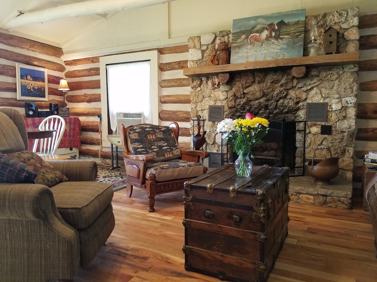 A cozy living area is highlighted by a stone fireplace and wooden logs. A floral arrangement sits on a wooden trunk coffee table. Comfortable seating options include an upholstered armchair and a patterned sofa, with artwork displayed above the fireplace and natural light entering from a window.