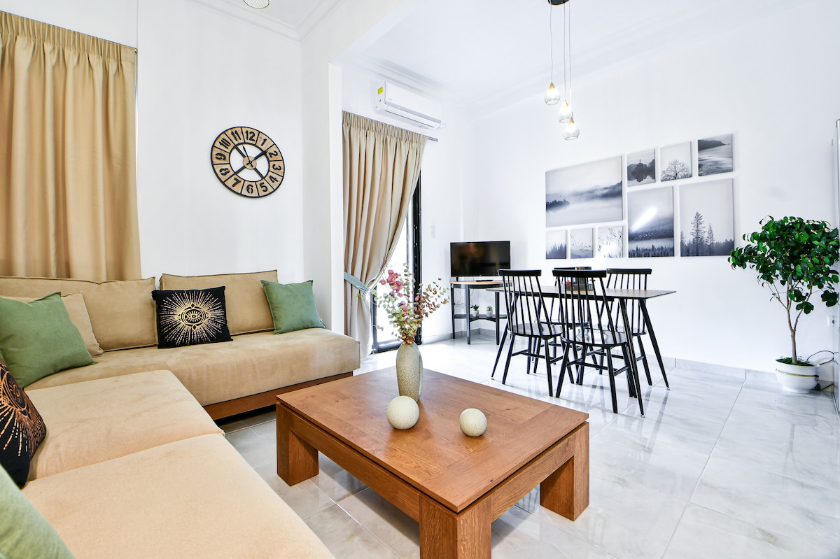 A modern living space features a beige sectional sofa adorned with decorative cushions. A wooden coffee table is complemented by subtle decor items. A dining table with black chairs is visible, and framed black-and-white photographs decorate the walls, enhancing the light-filled atmosphere.