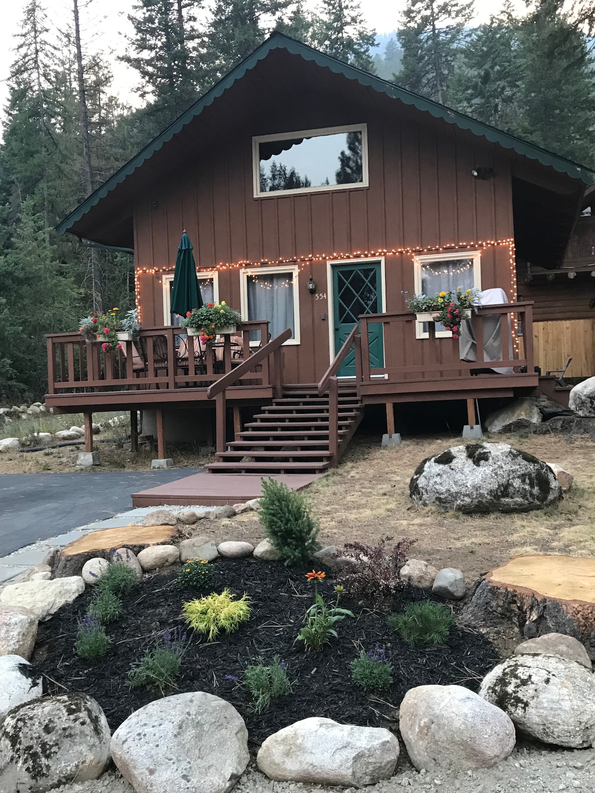 The chalet features a wooden exterior adorned with string lights, enhancing its charm. A welcoming front deck is accessed by steps, equipped with a green umbrella and flower planters. A landscaped area with rocks and plants frames the structure, contributing to a natural setting.
