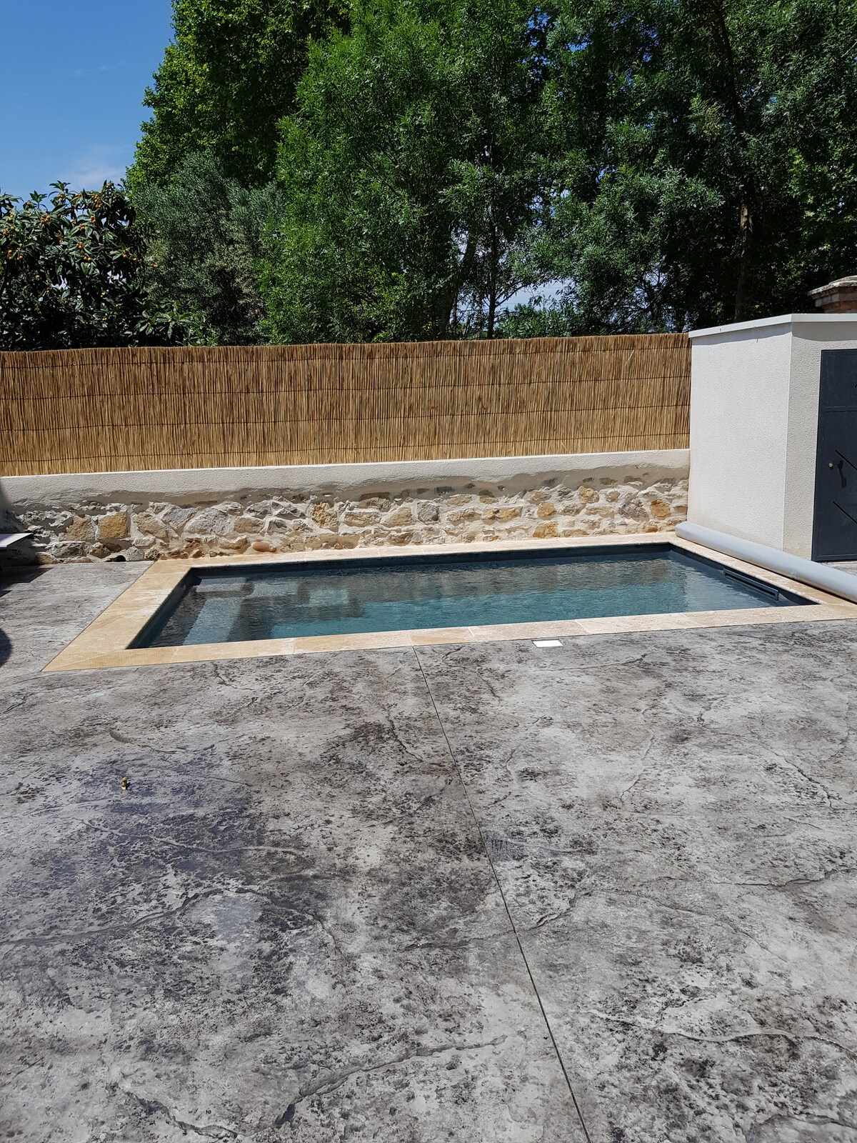 A small heated pool is displayed, surrounded by a stone patio. The space is bordered by a wooden fence and greenery, providing privacy. Soft shadows and sunlight reflect on the water's surface, creating a serene atmosphere.