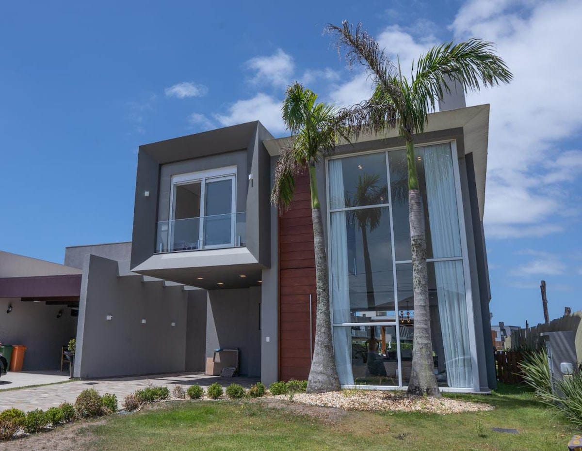 A modern house features large windows and a blend of textures on the exterior. Palm trees are positioned at the front, complementing the spacious lawn. The driveway is visible, leading to an outdoor area, enhancing the inviting atmosphere of the property.