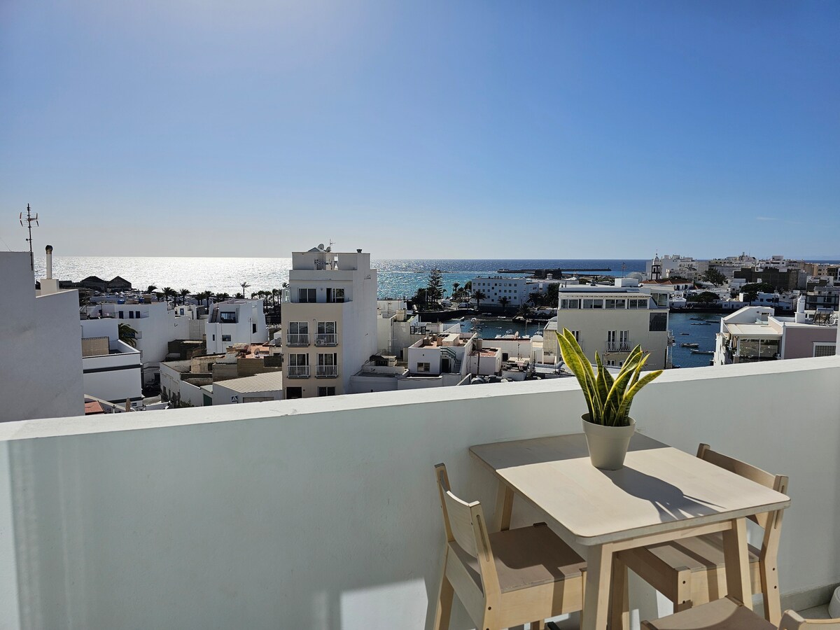 A terrace features a light wooden table with two matching chairs, complemented by a potted plant. The backdrop showcases a panoramic view of the coastline, with glistening water reflecting sunlight and a nearby harbor lined with white buildings.