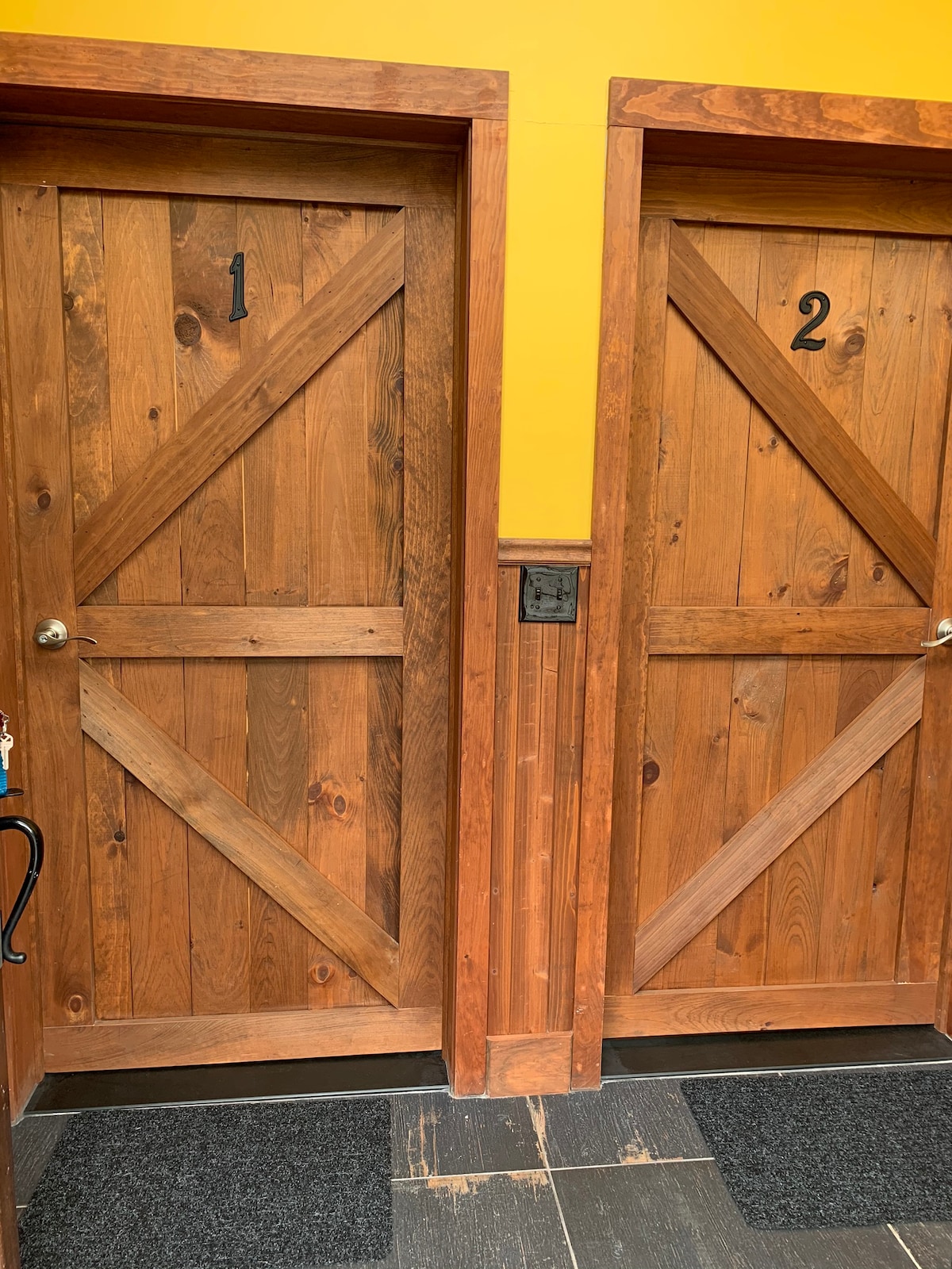 Two wooden doors are positioned side by side, featuring diagonal wood paneling and simple metal knobs. Each door bears a number, '1' and '2', prominently displayed. The entrance area is accented by dark flooring and a textured wall in a warm yellow hue.