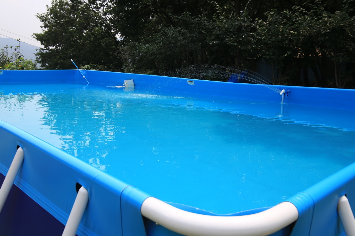 An above-ground swimming pool is visible, featuring clear blue water reflecting the surrounding greenery. The pool's walls are made of sturdy plastic, and water is being circulated from the built-in filtration system. The overall setting offers a refreshing escape in a natural environment.
