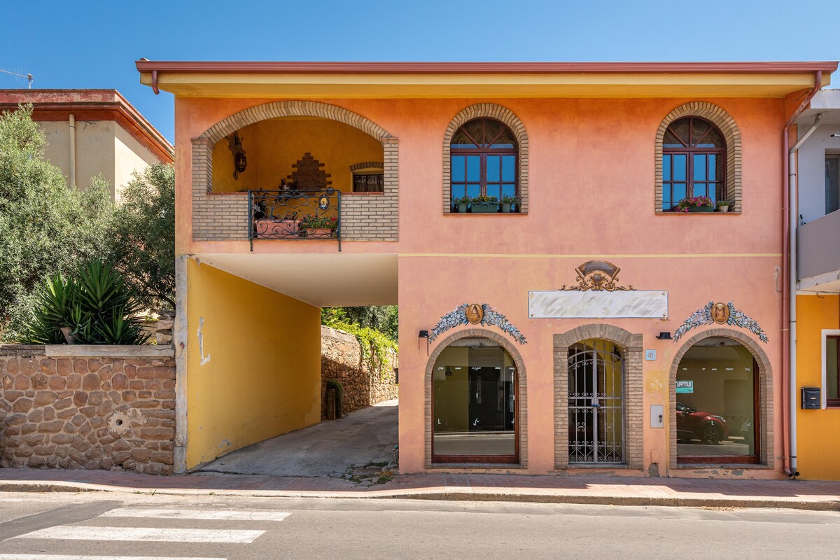 The exterior of the holiday home features a vibrant yellow facade with decorative stone accents. Arched windows allow natural light to fill the space, while the ground level offers an open area for parking. Lush greenery and Mediterranean plants surround the entrance.