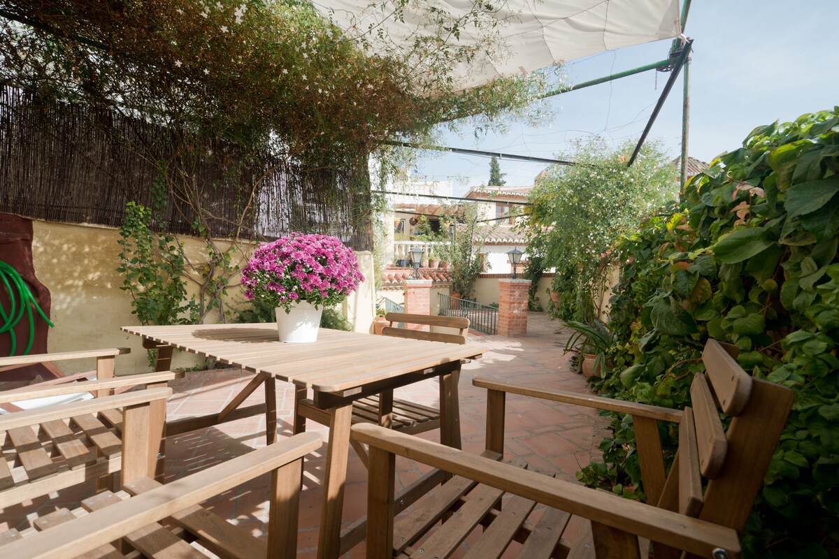 An inviting patio space is visible, featuring a wooden table surrounded by several chairs. A large pot filled with vibrant purple flowers serves as a focal point, complemented by green plants in the background. Shade is provided by a canopy above.