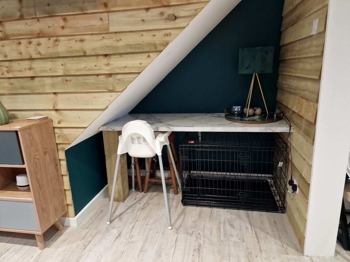 A small workspace is situated beneath a sloped ceiling, featuring a marble-top desk supported by wooden legs. A white high chair is positioned at one end, while a black pet crate is discreetly placed underneath the desk. The wall showcases natural wood paneling and a dark green accent section.