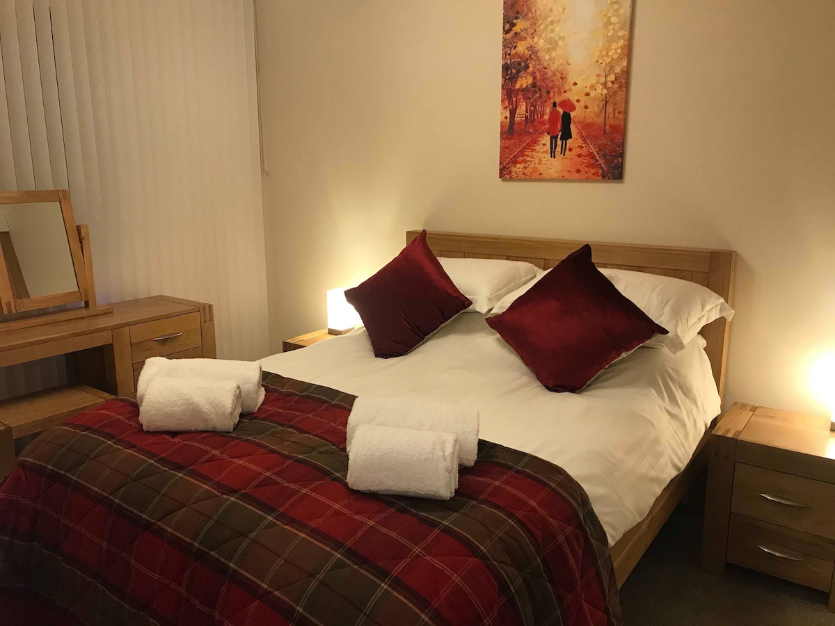 A well-appointed bedroom features a spacious bed dressed in white linens and topped with decorative red pillows. A warm plaid throw adds color at the foot of the bed. Two bedside tables, each with a lamp, flank the bed, while a framed artwork depicting a serene outdoor scene decorates the wall.