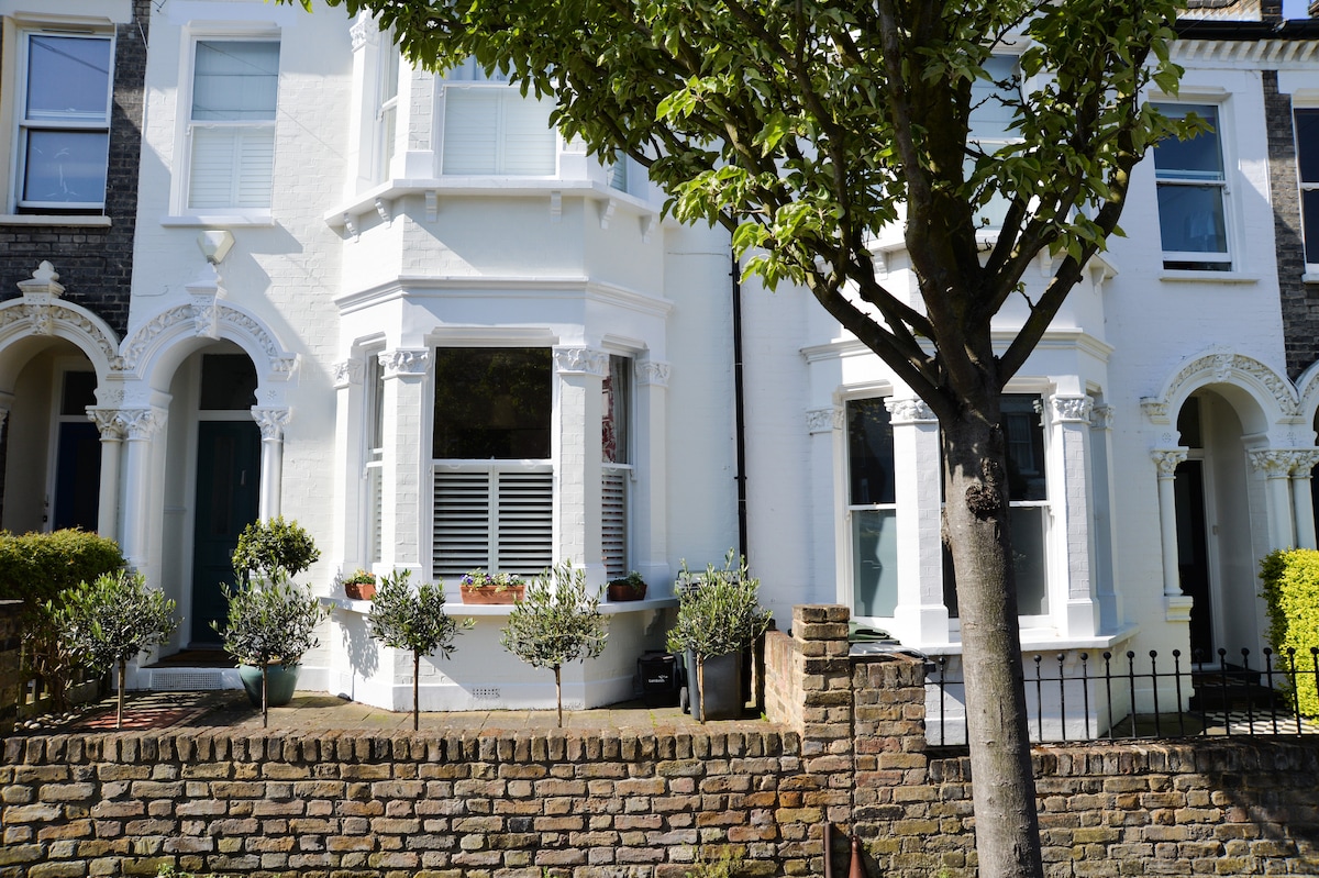 A charming Victorian terrace house features a vibrant white facade with decorative architectural details. Potted plants line the window ledge, and a small tree provides shade in front of the property. A low brick wall serves as a boundary, enhancing the welcoming appearance.