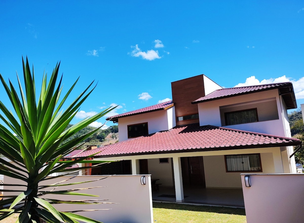 The exterior of a two-story house is shown, featuring a red-tiled roof and white walls. A decorative palm plant is positioned in the foreground. Large windows are visible, welcoming natural light, while the surrounding area is characterized by a well-maintained lawn and clear blue sky.