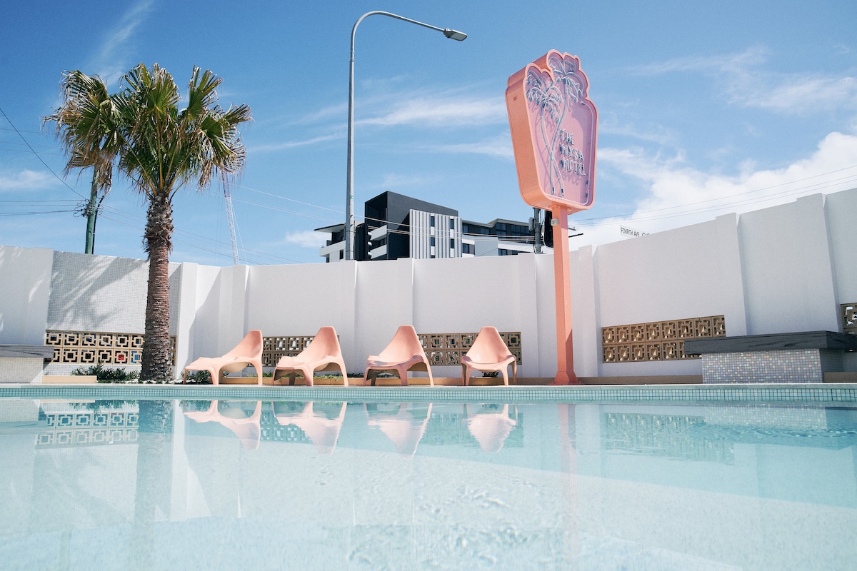 A serene view of a refreshing pool, surrounded by four stylish lounge chairs. A palm tree adds a tropical touch, while a retro sign stands prominently in the background against a clear blue sky, enhancing the character of the space.
