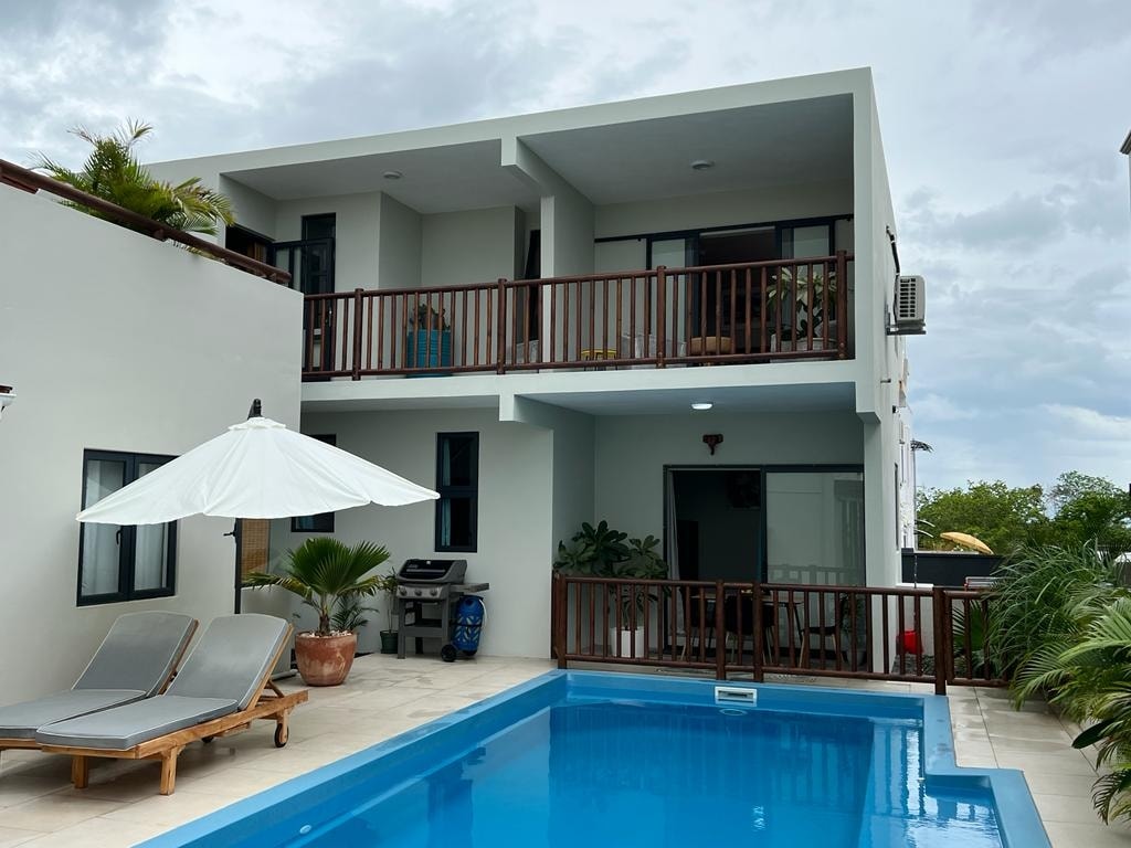 The exterior view captures a modern two-story house with wooden balconies and a spacious patio area. A salt-water pool reflects the surrounding greenery, accompanied by comfortable lounge chairs and a large umbrella. The residence is complemented by tropical plants, enhancing the serene environment.