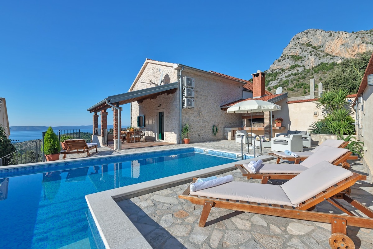 An outdoor area featuring a refreshing pool surrounded by sun loungers. A covered patio is visible, equipped with a dining table and lounge seating, backed by stone walls. The scenic view includes distant mountains and the sea, presenting a tranquil environment.