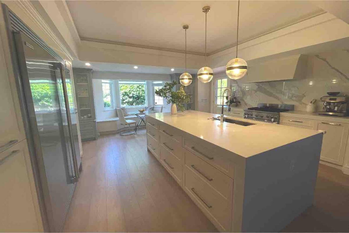 A spacious kitchen featuring a large island with modern light fixtures above. Stainless steel appliances are visible, along with ample cabinetry. Natural light fills the room through large windows, which overlook a cozy seating area with a small table and chairs.