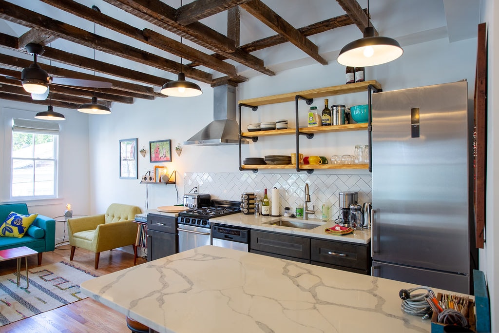 The kitchen features a modern design with dark cabinetry and a marble countertop. Open shelving displays dishes and kitchenware. Industrial-style pendant lights hang from exposed wooden beams. A stainless steel refrigerator and gas stove are visible, along with bright seating in the adjoining living area.