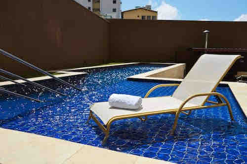 A tranquil swimming pool area features a tiled blue surface, complemented by a comfortable lounge chair positioned alongside. A white rolled towel rests atop the chair, with a gentle spray of water from the pool's edge, inviting relaxation under the clear sky.