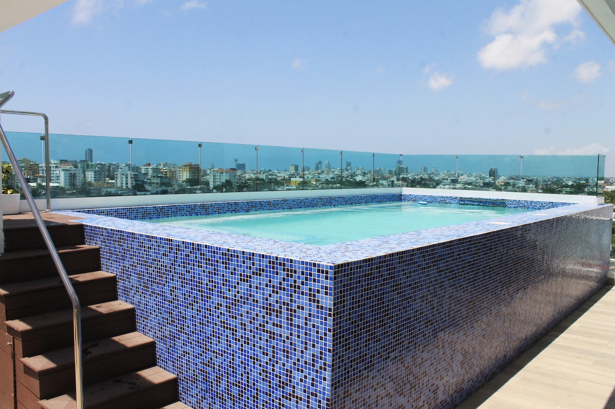 A rooftop pool featuring mosaic tiles in varying shades of blue is highlighted. The pool area offers a panoramic view of the surrounding cityscape, with a clear blue sky above. A wooden staircase leads up to the pool, enhancing accessibility.
