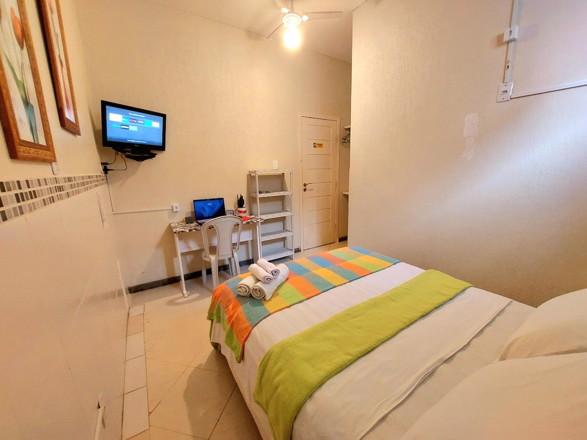 The room features a spacious bed with a bright green and blue striped blanket. A small TV is mounted on the wall, and a desk is positioned nearby, accompanied by a white chair. Towels are neatly arranged on the bed, with soft lighting illuminating the area.