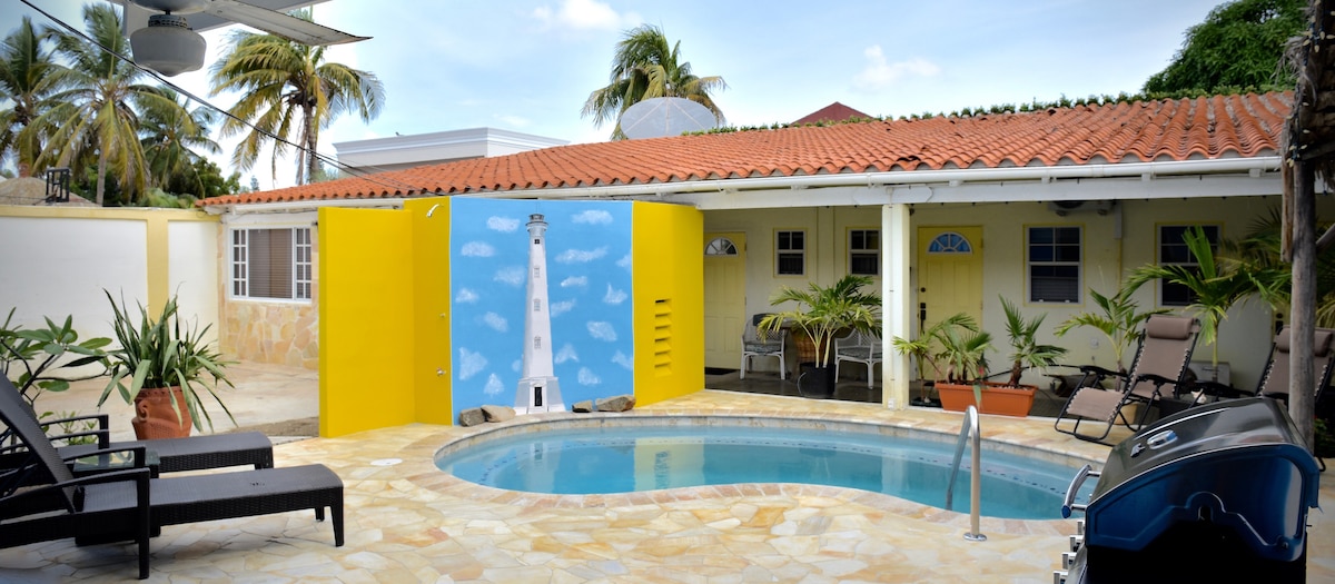 A pool area is surrounded by lounge chairs and tropical plants. A large mural of a lighthouse adds a vibrant touch to the yellow wall adjacent to the pool. The outdoor space is bathed in natural light, with a clear sky visible above and a tiled patio providing ample lounging space.