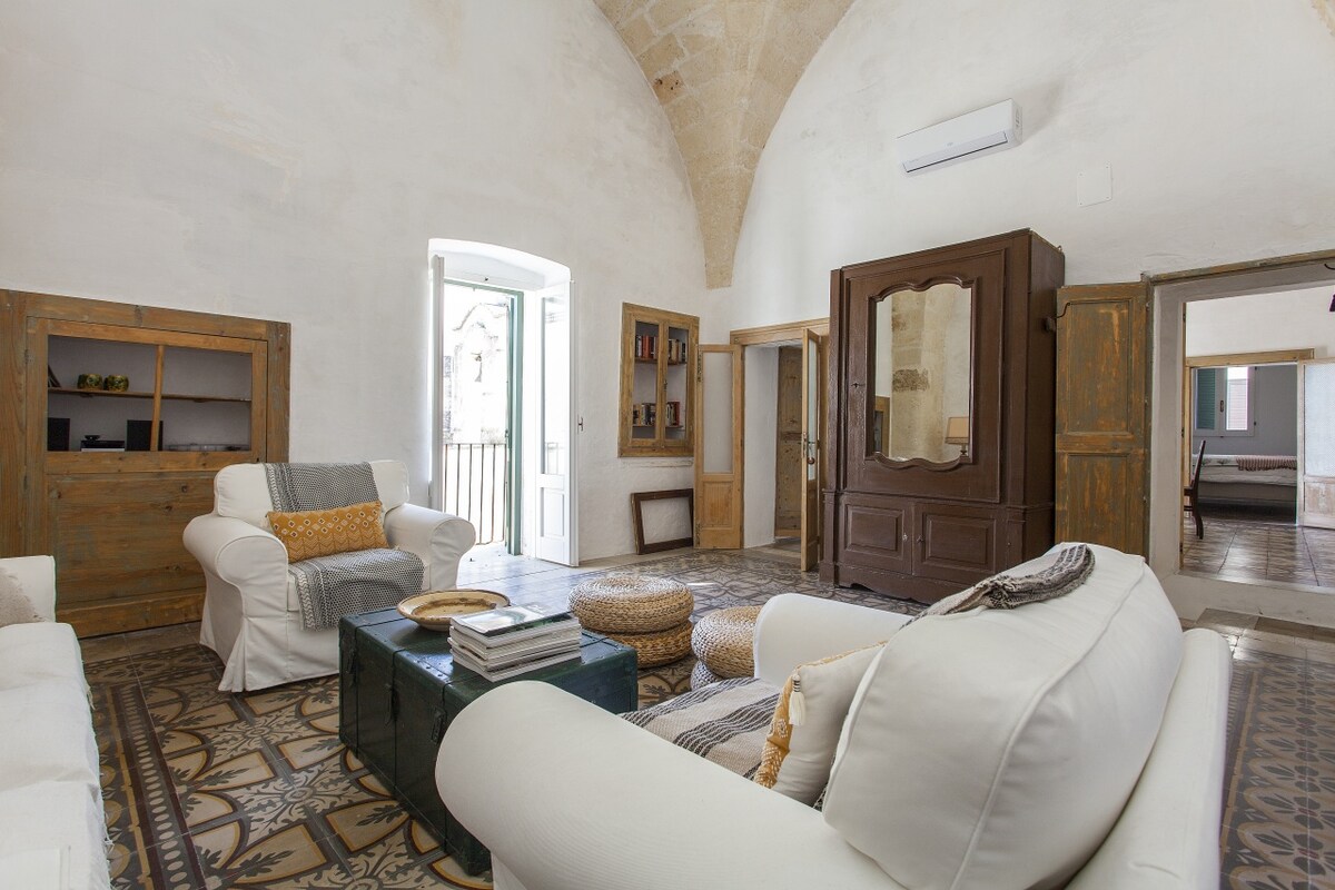 The living room features vaulted ceilings and comfortable seating, highlighted by a fold-out double sofa bed. A cozy coffee table is surrounded by poufs. Light streams through the open balcony door, and wooden cabinetry adds to the inviting ambiance of the space.