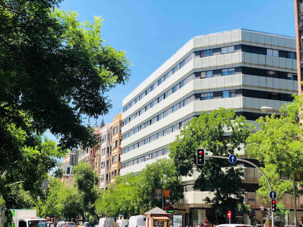 The exterior of a modern building is captured under a clear blue sky, bordered by lush green trees. A bustling street is visible below, with vehicles and a traffic signal marking the lively urban environment of the area. The contemporary design features large windows and a glass facade.