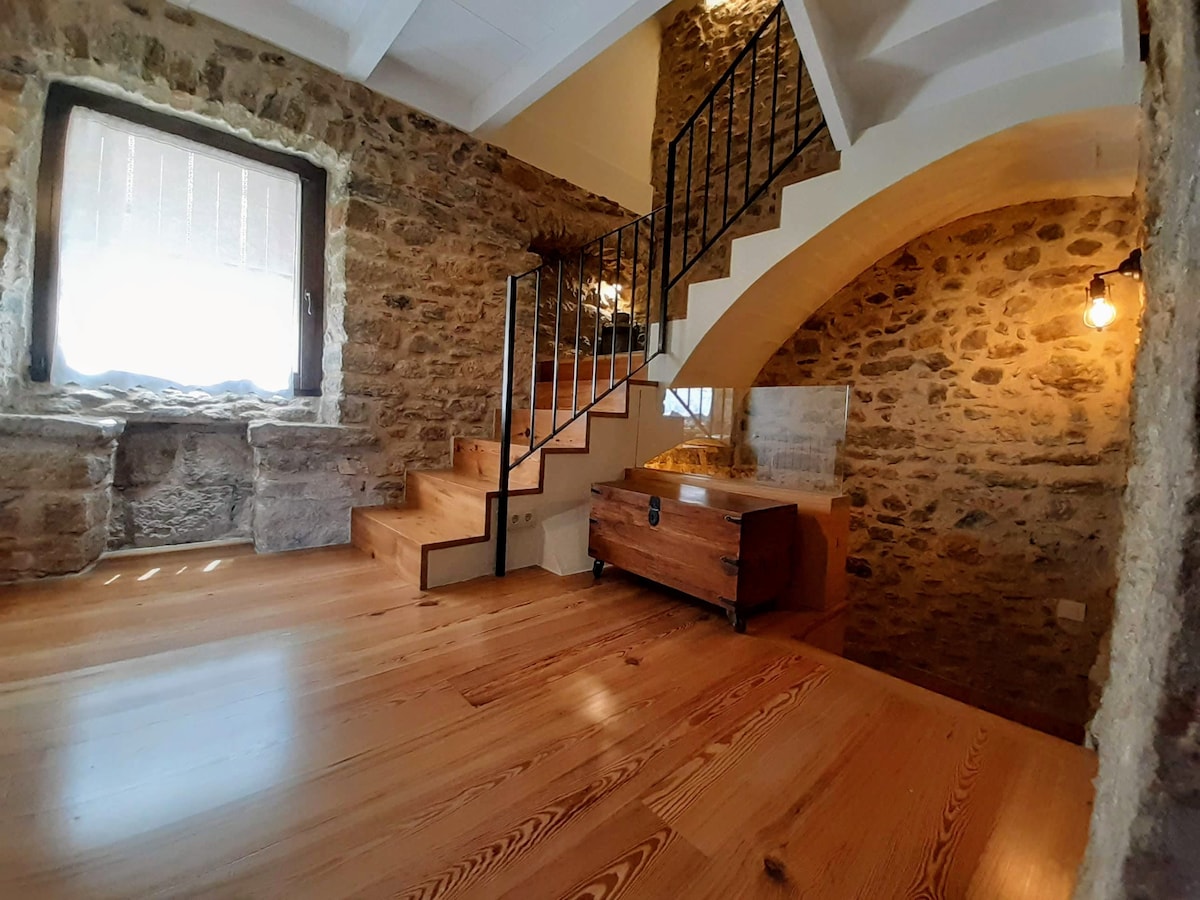 A bright interior space is showcased with warm wooden flooring and exposed stone walls. A staircase with a metal railing curves upward, leading to additional areas. Natural light filters through a window, creating an inviting atmosphere. A wooden chest is positioned against the wall.