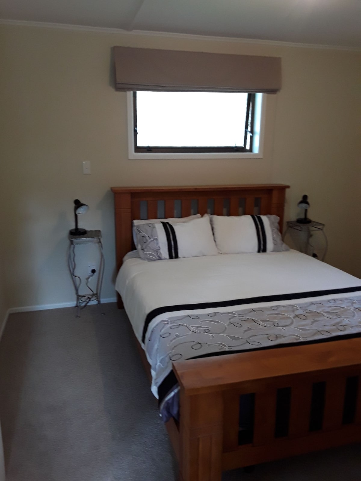 A separate bedroom features a wooden bed with a light-colored bedding set, complemented by decorative pillows. Two bedside tables with lamps are positioned on either side of the bed. A window allows natural light, and neutral-colored walls enhance the simplicity of the space.