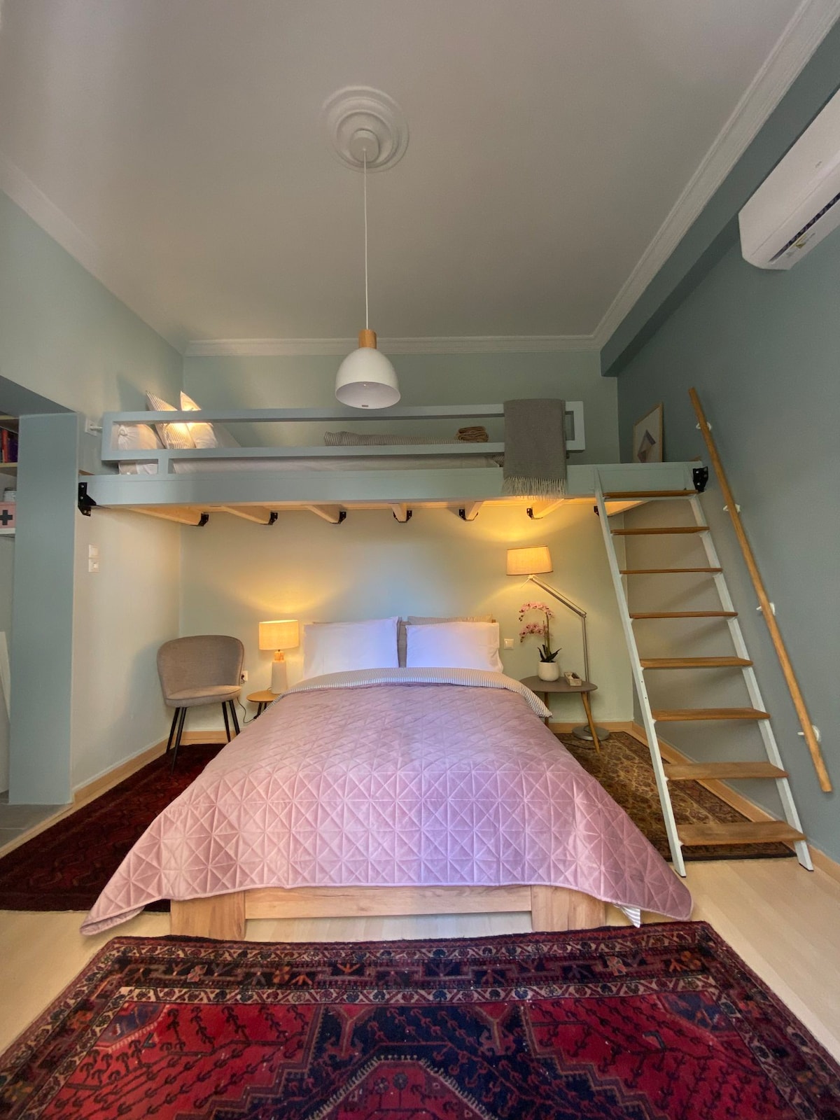 A cozy sleeping area is highlighted by a spacious bed covered with a light pink quilt. A ladder leads to a lofted area above. Soft lighting emanates from bedside lamps, and a comfortable chair is positioned nearby. Rich rugs adorn the floor, complementing the gentle blue walls.