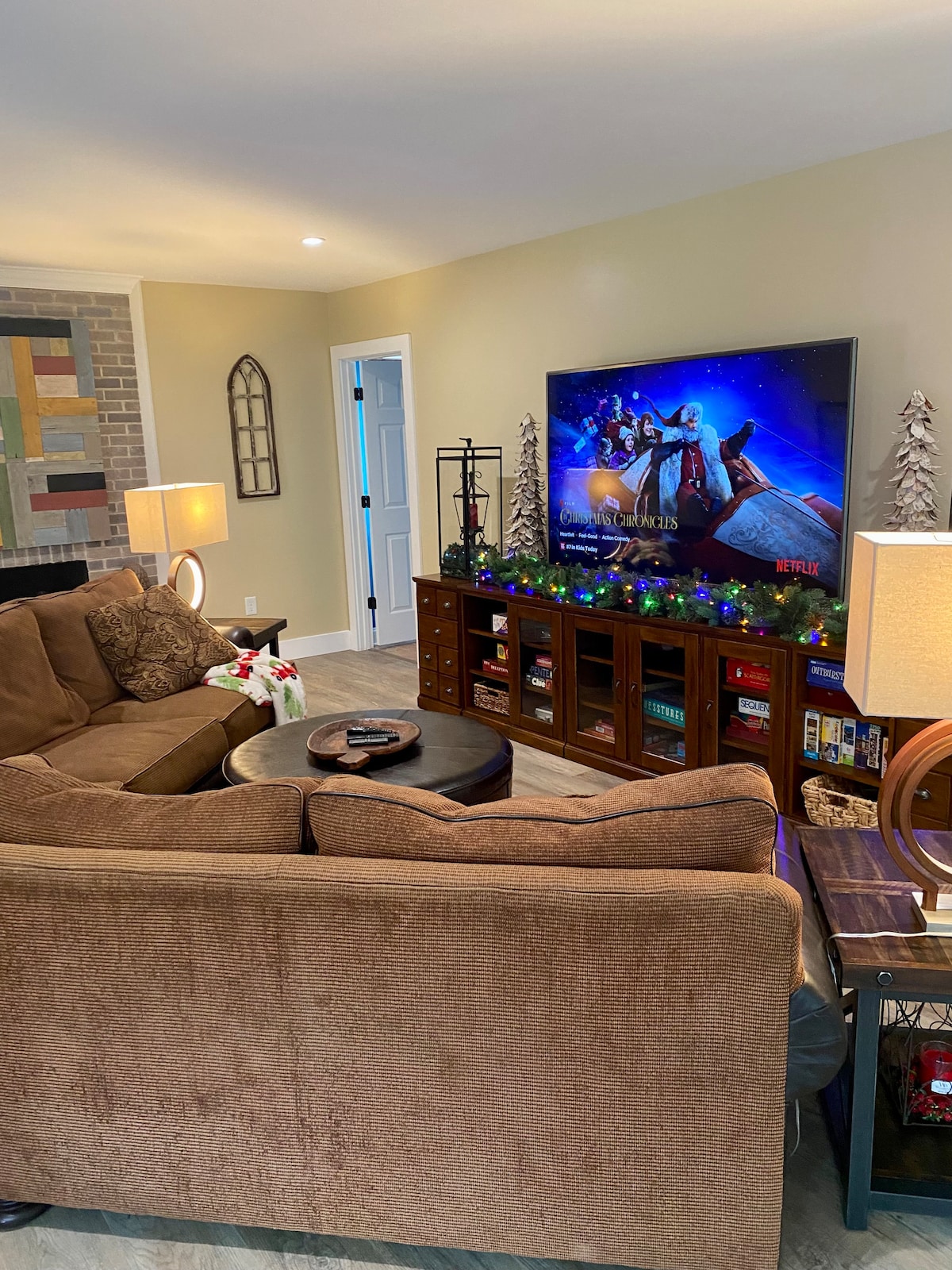 A cozy living area features a spacious sectional sofa, inviting relaxation. A large Smart TV is mounted on the wall, displaying colorful content. Two decorative lamps provide soft lighting, while a console table displays books and decorative items. The neutral walls create a warm backdrop.