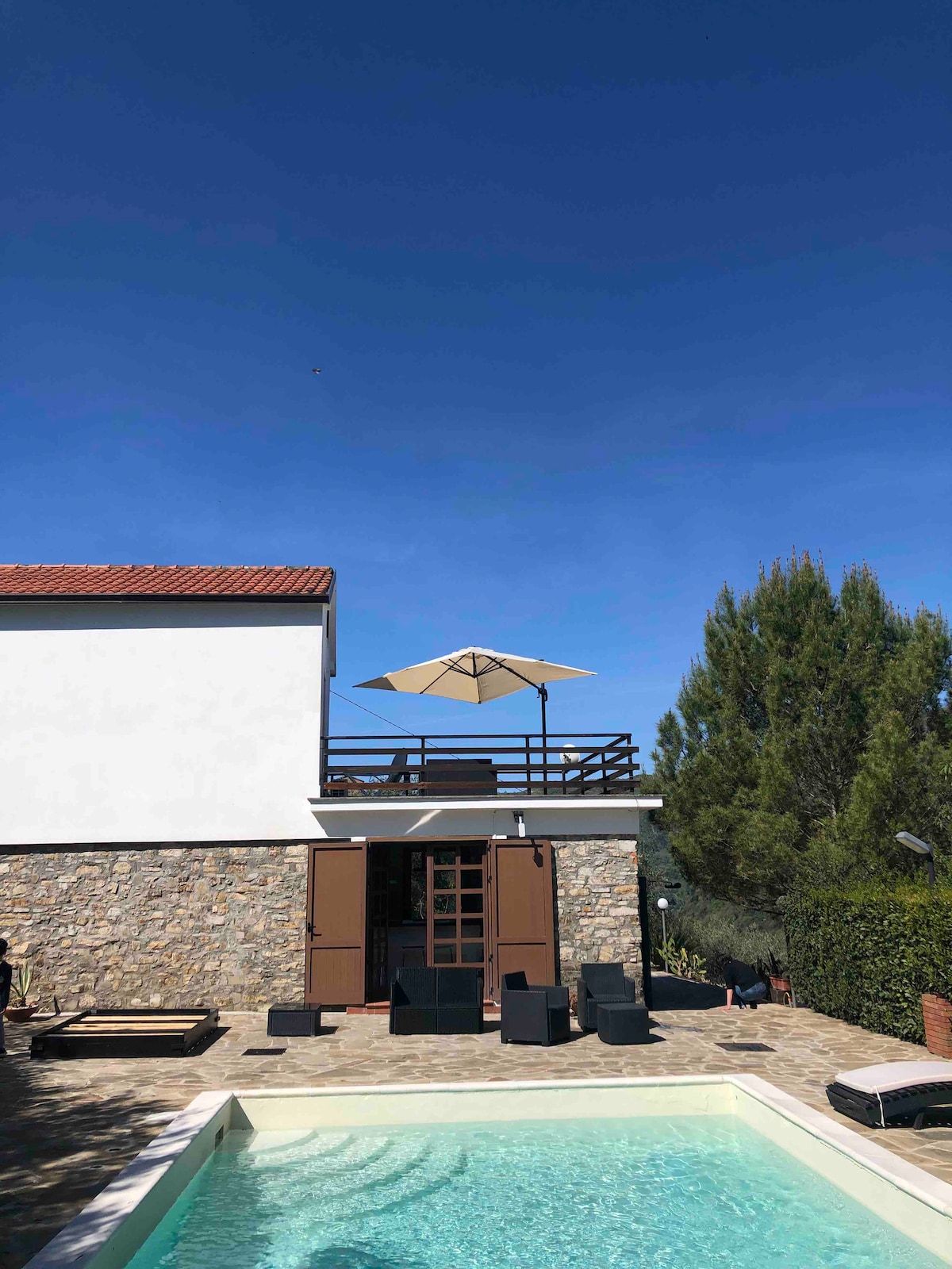A two-story country house with a stone exterior is depicted, featuring large open doors leading to a terrace. A sun umbrella provides shade over the seating area near the private pool. Lush greenery surrounds the space, complemented by a clear blue sky.