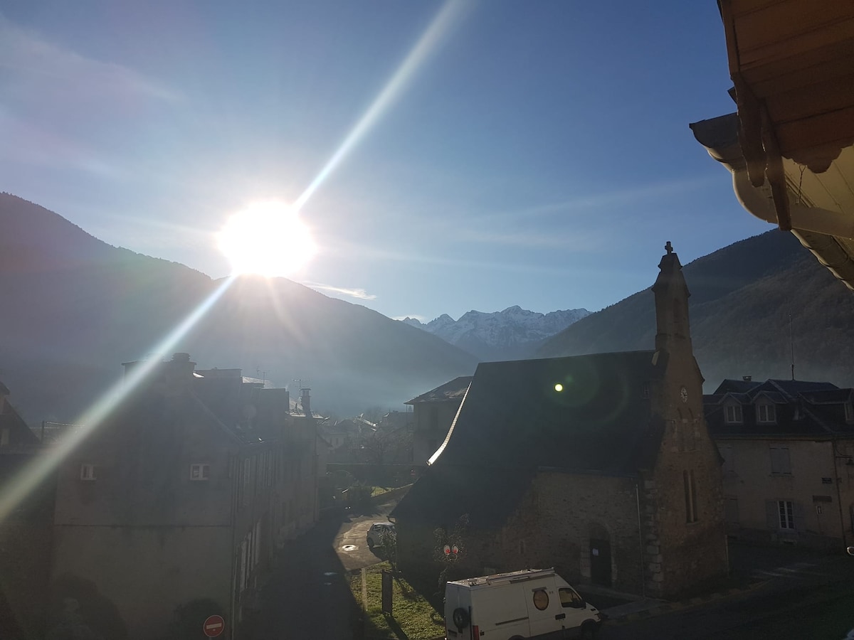 A bright sun illuminates the mountains in the background, with a charming village scene in the foreground. The image captures a church with a stone facade, nestled among low buildings, surrounded by the serene ambiance of a misty morning.