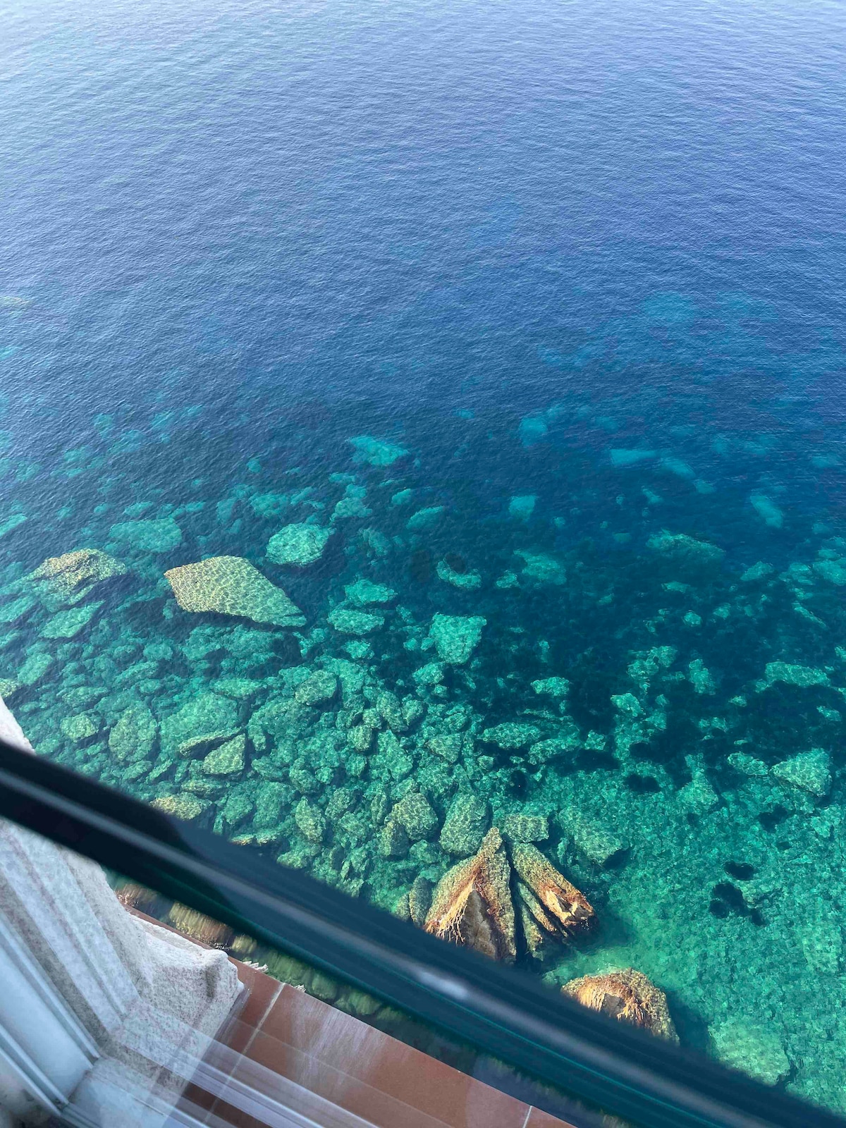 The image captures a view of the clear blue ocean from a high vantage point. Beneath the surface, various natural formations can be seen, including rocks and coral, creating a colorful underwater landscape. The sunlight reflects delicately on the water's surface.