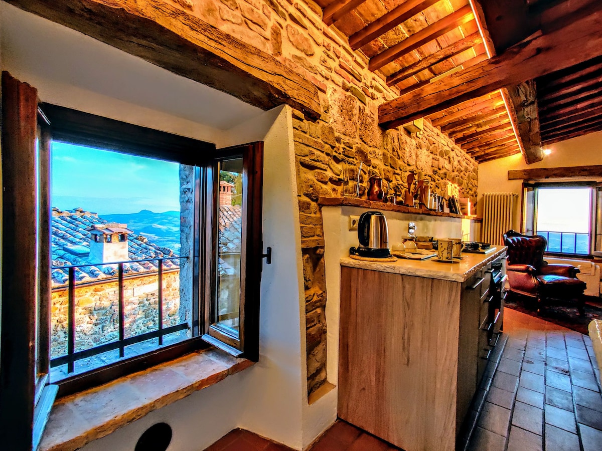 The kitchen area features exposed stone walls and wooden beams, providing a rustic charm. A large window overlooks the surrounding landscape, while countertops are equipped with modern appliances. A cozy armchair and warm lighting create an inviting atmosphere.