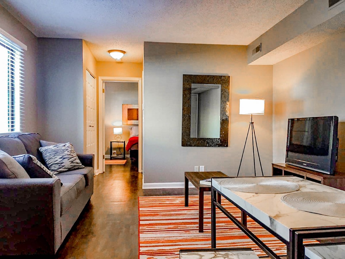A spacious living area features a comfortable grey sofa paired with a decorative blue and white cushion. A modern coffee table with a marble top sits on a striped area rug, while a tall lamp and a large mirror provide additional light and visual interest. A doorway leads to another room.