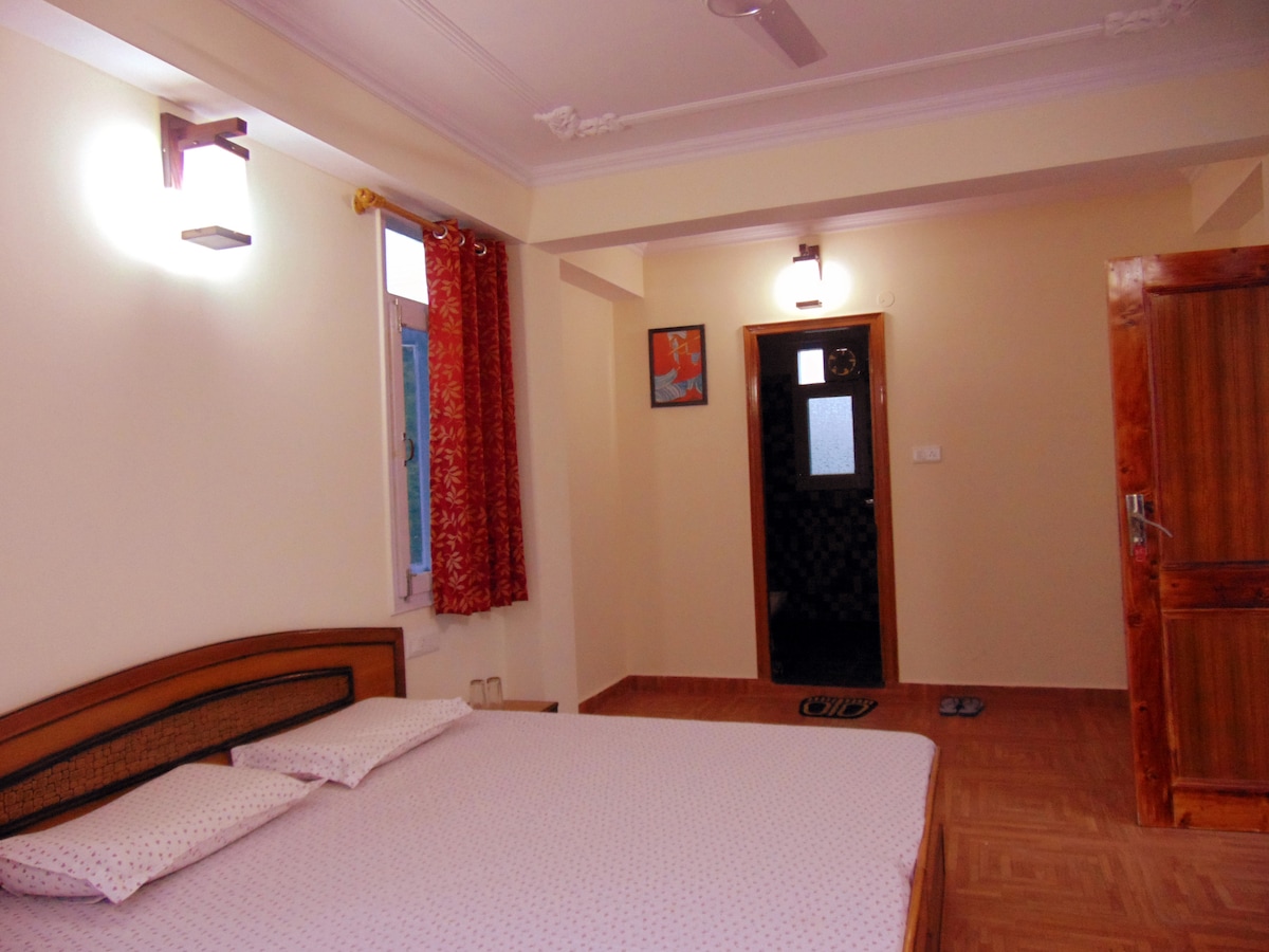 A well-furnished bedroom features a spacious bed with a subtle patterned mattress. Natural light enters through a window adorned with patterned curtains, while modern light fixtures add a soft glow. A wooden door leads to the hallway, and a piece of wall art decorates the room.