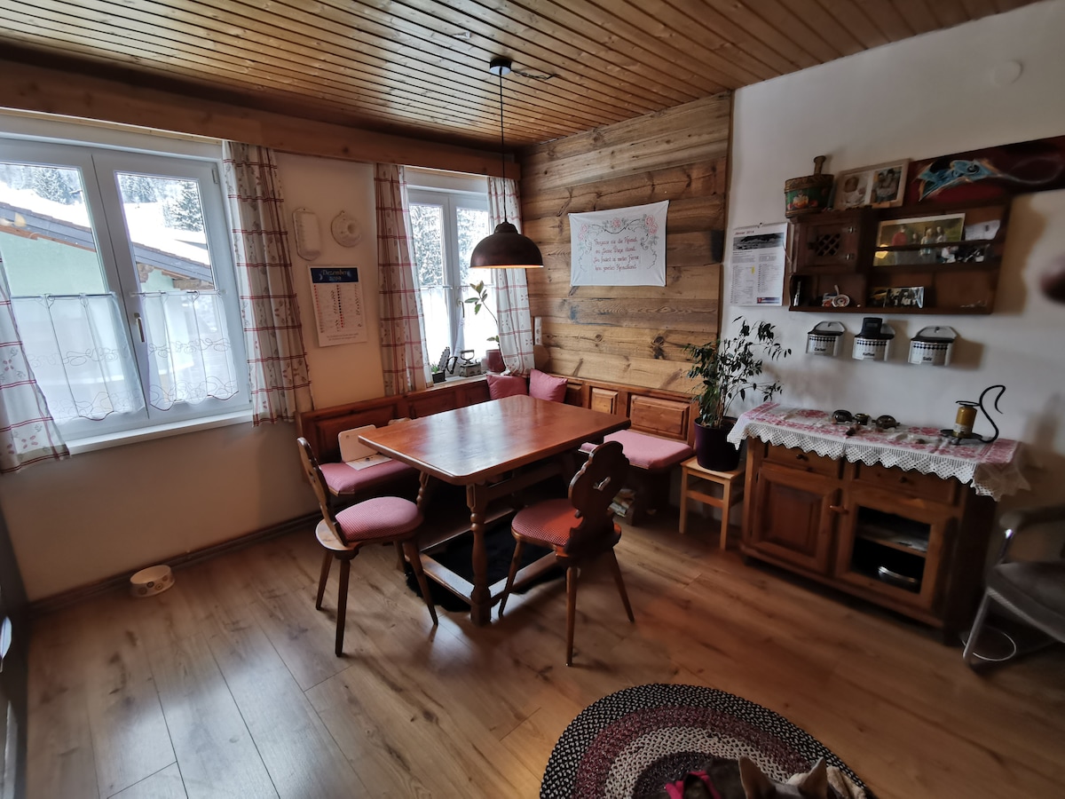 A rustic dining area features a wooden table surrounded by chairs and a cozy bench, with soft pink cushions. Large windows allow light to fill the room, offering views of the outdoors. A wooden shelf displays decorative items and plants, enhancing the inviting atmosphere.