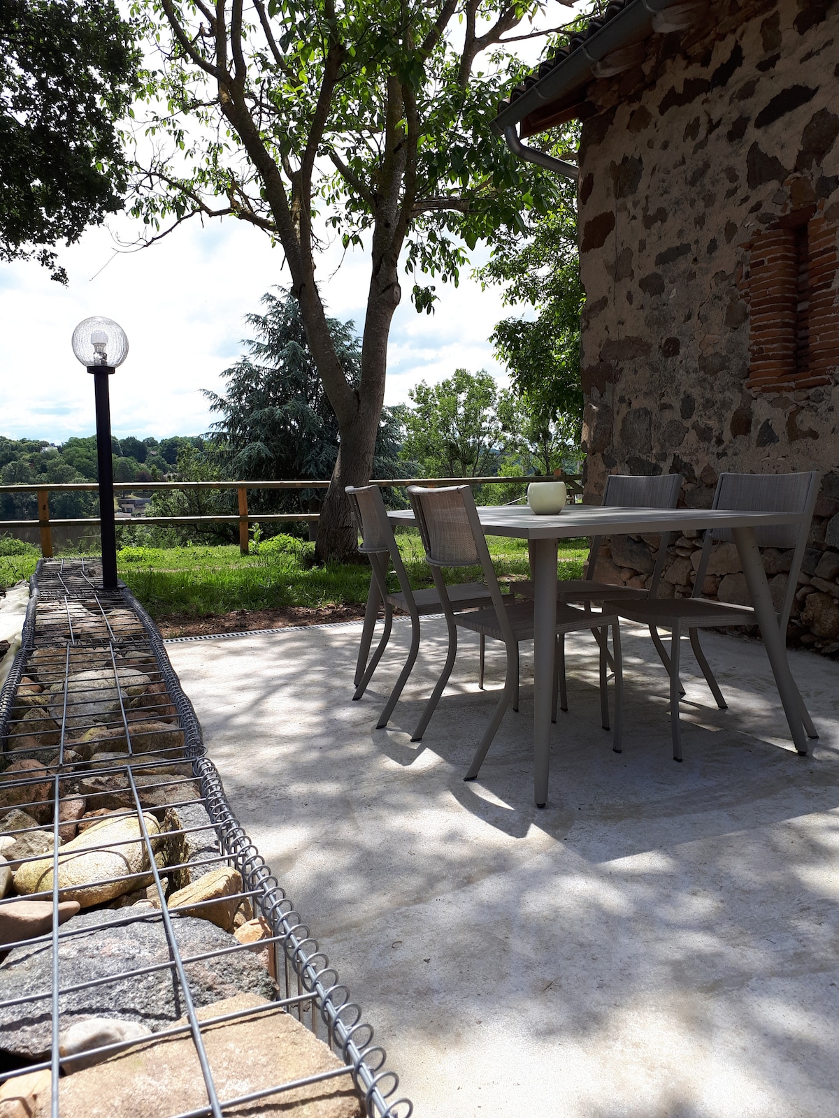 An outdoor seating area is presented with a light gray table and four matching chairs placed on a concrete patio. A stone border is visible along the edge, and a round lamp stands nearby, casting light across the inviting space surrounded by greenery.