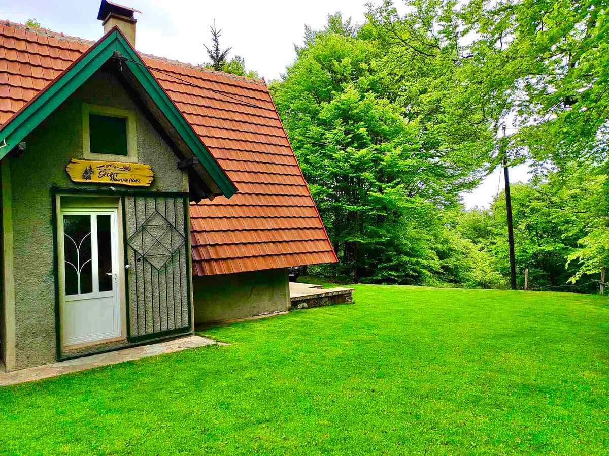 A charming cottage exterior is highlighted by a sloped roof with rustic red tiles. A well-kept lawn extends in front, surrounded by lush greenery. The entrance, framed by a decorative door, features a simple porch area with stone steps leading to the ground.