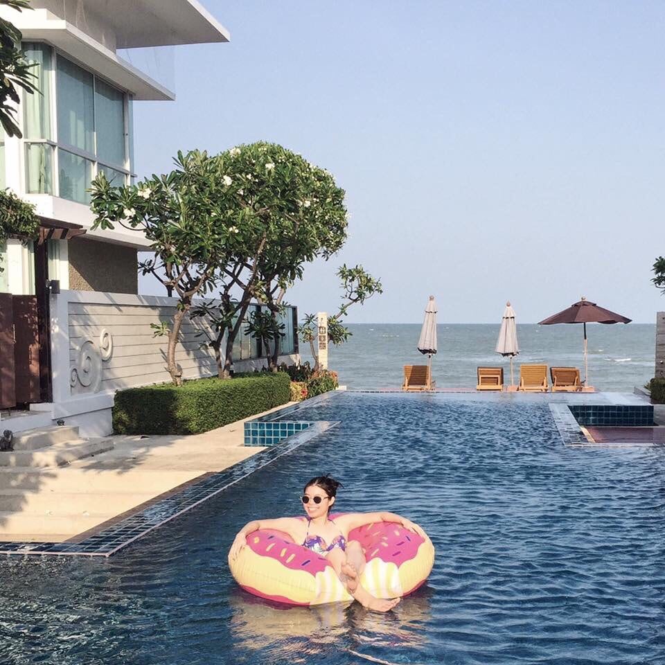A serene pool area is surrounded by tropical plants and modern architecture. A colorful inflatable float rests in the water, while sun loungers with umbrellas are visible in the background, facing the beach. The clear sky enhances the tranquil atmosphere of the setting.