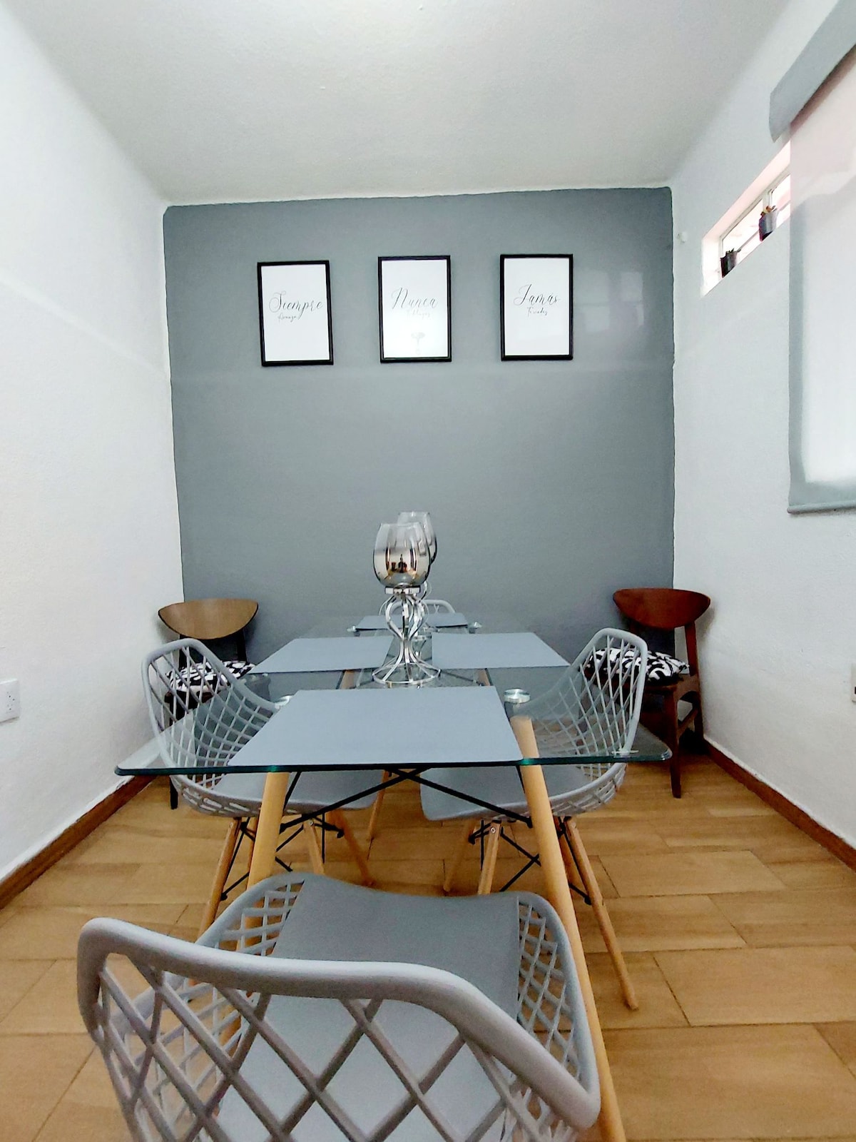 A dining area features a modern glass table surrounded by four sleek chairs. Two art pieces are displayed on the gray wall, while the floor showcases light-colored tiles. Natural light filters in through a window adorned with a light grey shade.
