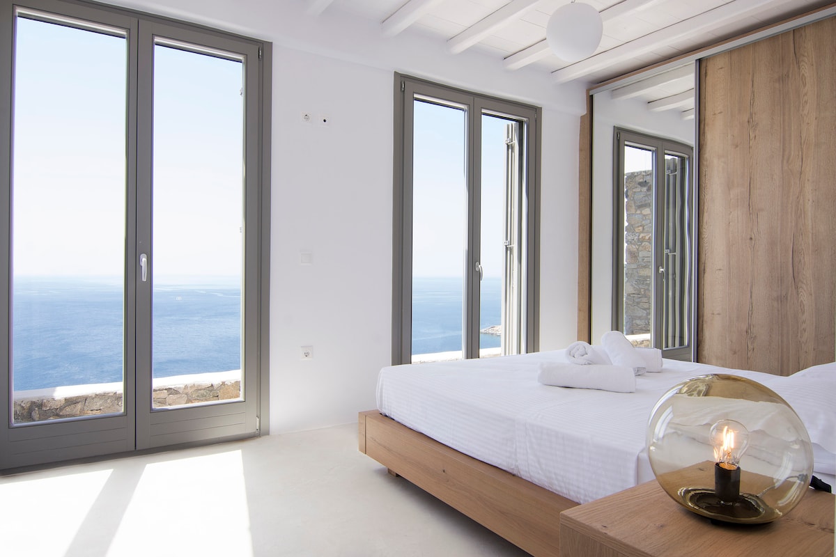 A bright master bedroom features large glass doors opening to a panoramic sea view. Light walls and a minimalistic design create an airy space, complemented by a wooden bed and a decorative lamp. Fresh linens are arranged neatly on the bed, enhancing the room's serene ambiance.