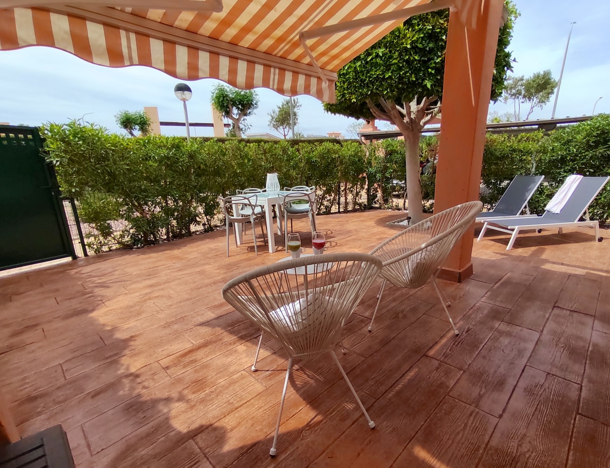 A private terrace features a shaded area with two white chairs and a glass table set for four. Lush greenery surrounds the space, providing privacy, while sun loungers await for relaxation. A retractable awning offers additional shade against sunlight.