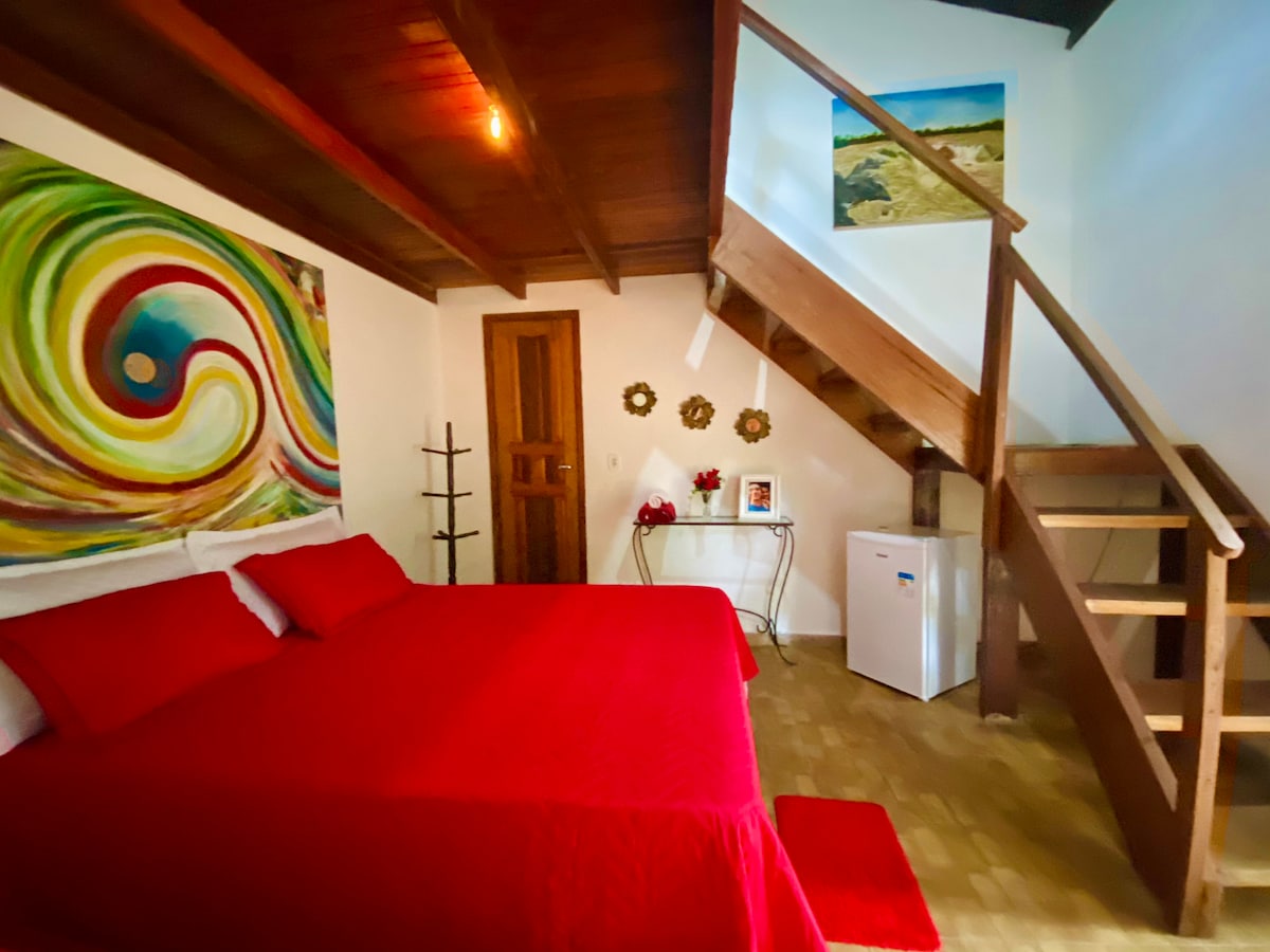A spacious sleeping area with a red bedspread covers a large bed. Colorful artwork adorns the wall, complementing the wood-paneled ceiling. A staircase leads to the mezzanine, while a small fridge is positioned in the corner, enhancing the room's practicality.