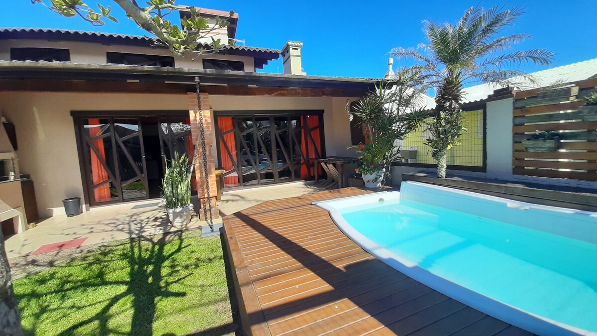 An outdoor area features a wooden deck surrounding a small pool. Lush greenery and potted plants provide a natural touch. Large glass doors lead into the interior, framed by vibrant red curtains, while the clear blue sky enhances the inviting atmosphere.