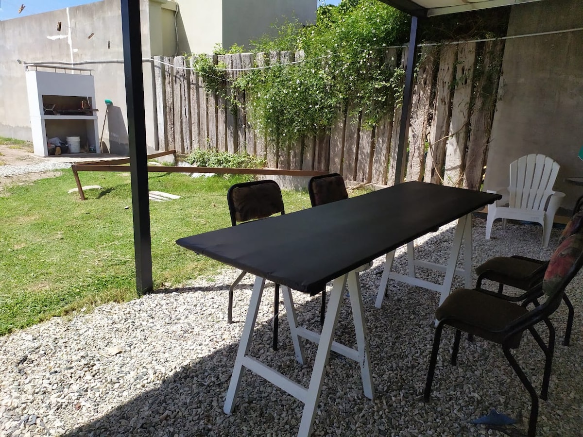 An outdoor dining area is present, featuring a long black table surrounded by chairs on a gravel surface. Lush greenery and a wooden fence are visible in the background, contributing to a serene environment. A barbecue grill is located to the left, offering additional outdoor cooking options.