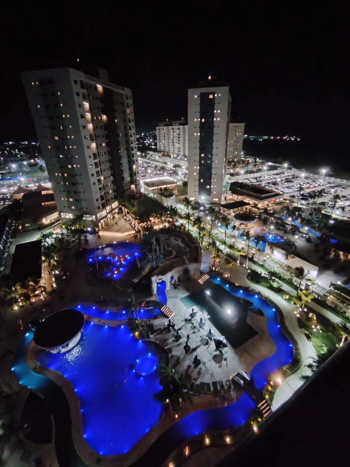 An aerial view captures the resort at night, showcasing illuminated pools with varying shapes and depths. Surrounding the water features, palm trees are highlighted by soft lighting. Nearby buildings rise in the background, reflecting the vibrant atmosphere of the resort and its surroundings.