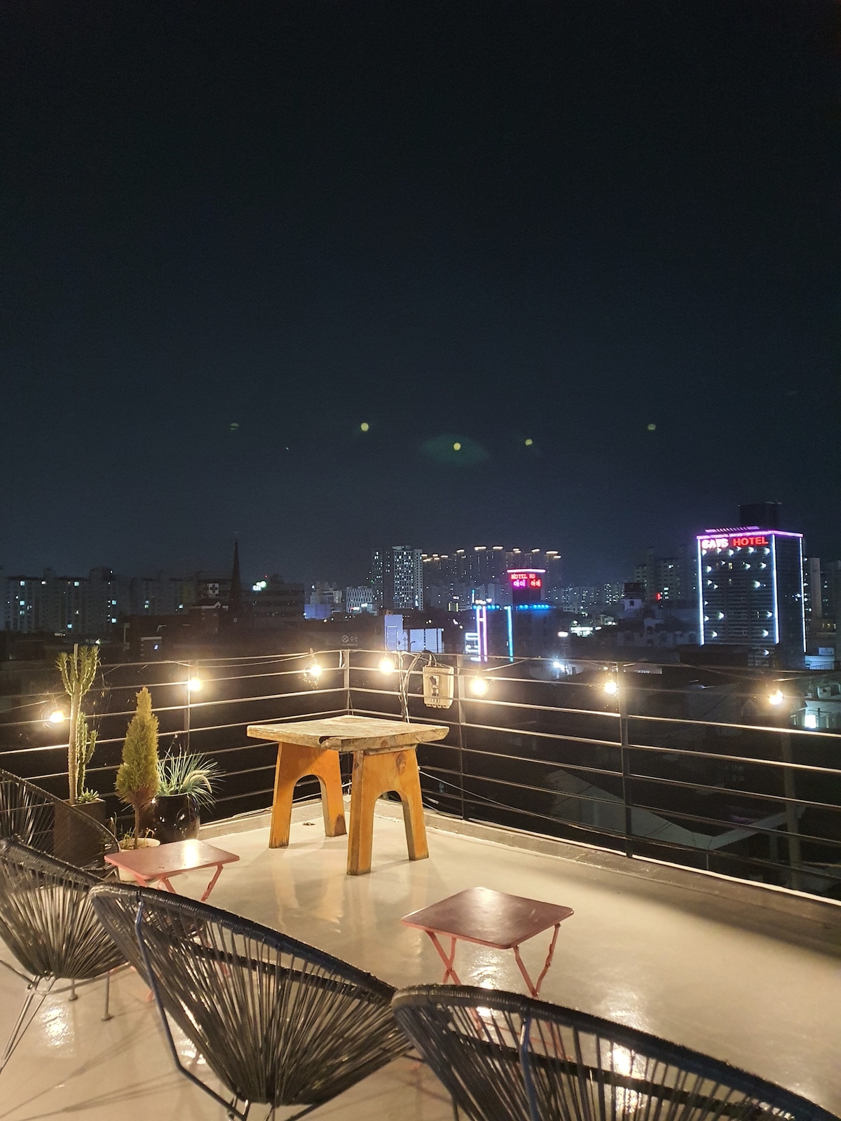 An outdoor rooftop space is illuminated under a starry night sky. A wooden table sits at the center, surrounded by black chairs. The city skyline, featuring various buildings and neon lights, is visible in the background, creating a vibrant atmosphere.